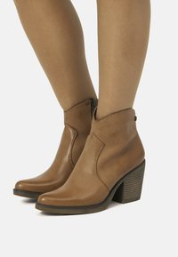 Botas de cuero marrón hasta el tobillo con puntera puntiaguda, detalles de costura y un tacón bajo en bloque. Cuentan con una cremallera lateral para facilitar su uso.