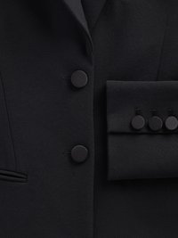 Veston noir avec des boutons ronds noirs, une poche poitrine unique et un tissu lisse et texturé avec une coupe cintrée.