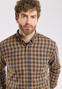 Homme avec des cheveux bouclés et une barbe, portant une chemise à carreaux marron et bleu marine à boutons, faisant face à l'avant sur un fond clair.