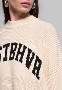 Beige gestrickter Pullover mit gerippter Struktur und schwarzen Buchstaben. Hoher Kragen, lockere Passform und kontrastierende Abschnitte auf der Brust.
