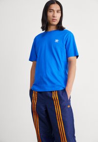 Blå bomulds t-shirt med rund halsudskæring og hvid Adidas-logo, parret med marineblå mønstrede bukser med orange striber og et accent-logo.