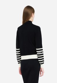 Cardigan nero con colletto alto, con accenti bianchi a righe sulle maniche e sull'orlo. Tessuto morbido con dettaglio del logo sulle maniche.