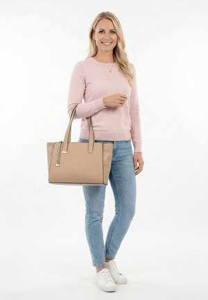 Kobieta w różowym swetrze i jasnoniebieskich dżinsach, trzymająca beżową torbę typu tote, stojąca i uśmiechająca się na białym tle.
