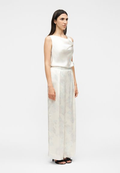 Vrouw staat, draagt een mouwloze witte satijnen top met gedrapeerde halslijn en hoge taille, wijd uitlopende bloemenbroek, gecombineerd met zwarte open-toe hakken.