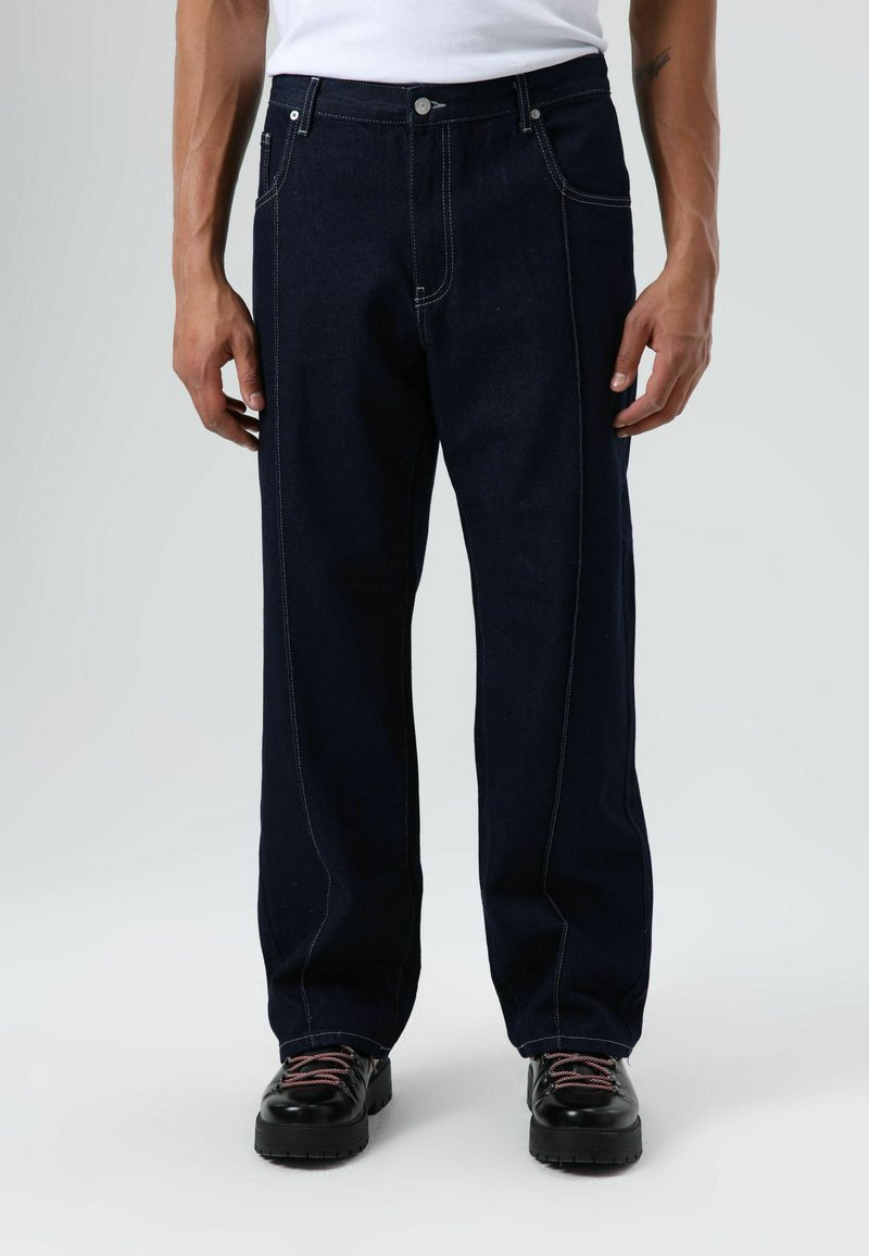 Donkerblauwe jeans met een rechte pijp, contrasterende stiknaden, vijf zakken en een knoopsluiting, gecombineerd met zwarte sneakers.