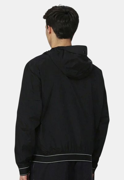 Uomo che indossa una giacca nera con cappuccio e dettagli bianchi, in piedi di spalle contro uno sfondo semplice.