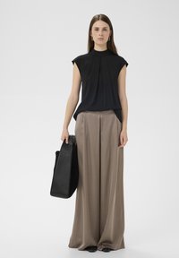 Fekete rövid ujjú blúz redőkkel, világosbarna, bőszárú nadrággal párosítva. A modell egy fekete bőrtáskát tart a kezében.