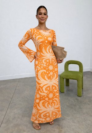 Naine seisab siseruumides, kandes pikk oranži kleidi beežide lillemustritega, hoides käes beeži õlakotti, rohelise moodsa tooli lähedal.