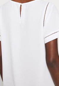 Blouse blanche avec fermeture à dos en forme de goutte, manches courtes et découpes décoratives le long des ourlets des manches. Tissu lisse et léger.