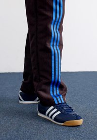 Mørkebrune træningsbukser med to blå striber på hver side, parret med marineblå sneakers med tre hvide striber og en gummisål.