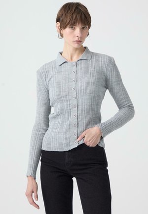 Grå ribbad kofta med krage, med knappslutning fram och långa ärmar. Bärs över mörka jeans.