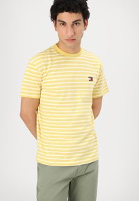 Tricou cu dungi galbene și albe, cu mâneci scurte, guler rotund și un mic logo pe pieptul stâng. Material moale, din bumbac.