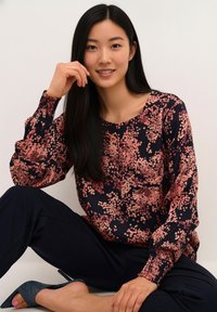 Blouse bleu marine à manches longues avec imprimé floral rose, encolure ronde et manches froncées, associée à un pantalon foncé et une chaussure grise à talon haut.