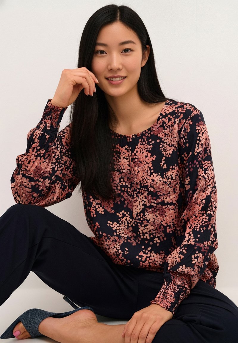 Blouse bleu marine à manches longues avec imprimé floral rose, encolure ronde et manches froncées, associée à un pantalon foncé et une chaussure grise à talon haut.