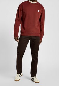 Bordowa bluza z okrągłym dekoltem z prążkowanymi mankietami i dołem, zestawiona z ciemnobrązowymi spodniami oraz białymi trampkami z czarnymi paskami.