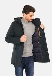 Cappotto impermeabile navy con cappuccio, dotato di chiusura con zip e bottoni a pressione, foderato in testa piuma di colore blu scuro, con tasca a contrasto arancione.