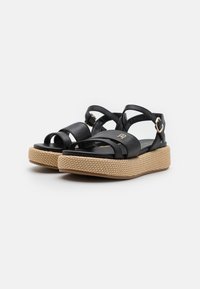 Black leather platform sandals with crisscross straps, a rounded toe, and a woven jute texture midsole. Features a metallic accent emblem.