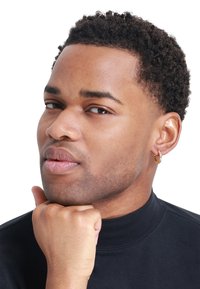 Boucle d'oreille en or avec un petit strass transparent incrusté. Le mannequin a les cheveux courts et texturés de couleur noire et porte une chemise noire à col haut.