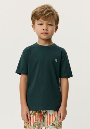 Młody chłopiec o jasnobrązowych włosach nosi ciemnozielony t-shirt z podwiniętymi rękawami i kolorowe, wzorzyste szorty, stoi na jednolitym tle.