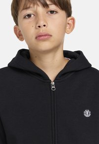 Sudadera con cremallera negra hecha de un tejido suave, con una cremallera plateada, que presenta un pequeño logo blanco en el área del pecho izquierdo.