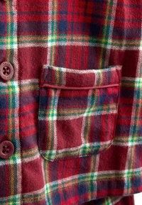 Czerwona i zielona koszula flanelowa w kratę z dwoma ciemnymi guzikami, kieszenią na piersi i miękką teksturą. Wzór zawiera białe i granatowe linie.