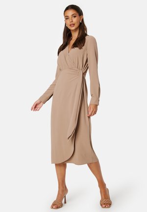 Wikkeljurk van lichtbeige stof met lange mouwen, een V-halslijn, zijstrikdetail en een vloeiende silhouet, gecombineerd met geweven haksandalen.