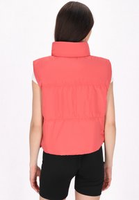 Gilet smanicato imbottito, di un vivace colore corallo, colletto alto, dettagli cuciti, texture liscia, indossato sopra una maglietta bianca e pantaloni corti neri.
