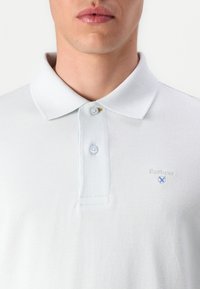 Ανοιχτό γκρι πόλο μπλουζάκι φτιαγμένο από υφασμάτινο υλικό, με κλασικό γιακά, δύο κουμπιά και ένα μικρό μπλε κεντημένο λογότυπο στο στήθος.