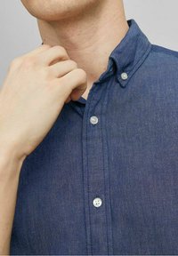 Košile na knoflíky z tmavě modré látky, s límcem, jemnou texturou a průhlednými plastovými knoflíky, ruka upravující oblast kolem krku.