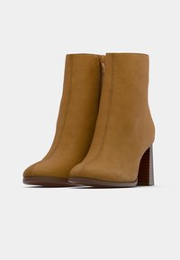 Barna színű velúr boka csizma keskenyített orral, magas tömbsarkúval és oldalsó cipzárral. A dizájn letisztult vonalakat és tónusos varrást tartalmaz.