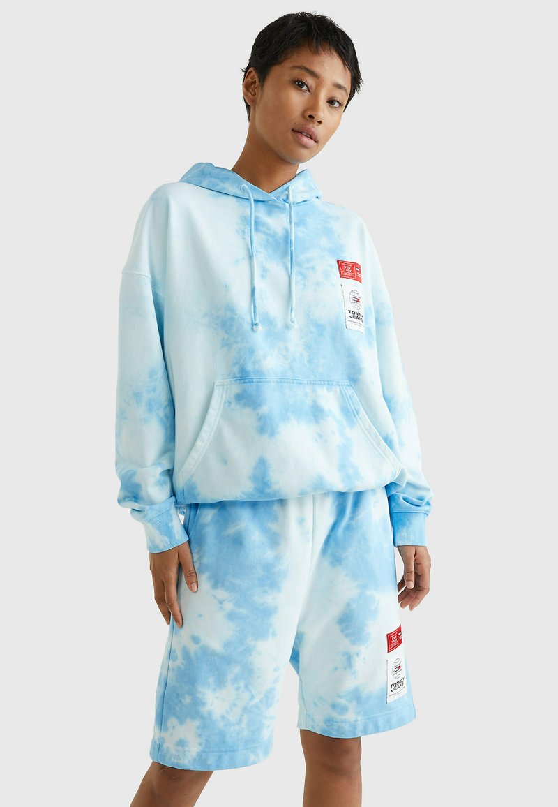 Ensemble hoodie et short en tie-dye bleu clair, fabriqué en tissu doux, avec une poche avant et deux étiquettes en haut à gauche du hoodie.