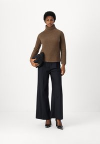 Brauner gerippter Rollkragenpullover, weite schwarze Hose und schwarze Absatzschuhe. Hält eine strukturierte schwarze Clutch. Einheitlicher weißer Hintergrund.