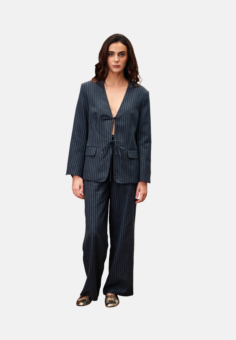 Frau in einem dunklen Nadelstreifenanzug mit einem Blazer mit Krawatte und weit geschnittenen Hosen, kombiniert mit metallischen Ballettflats, stehend vor einem weißen Hintergrund.