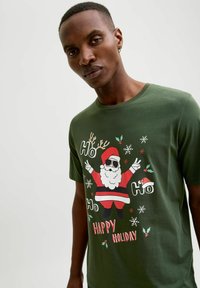 Camiseta de algodón verde con un diseño de Papá Noel usando gafas de sol, el texto "Ho Ho Ho" y "FELICES FIESTAS", junto con copos de nieve y acentos de acebo.