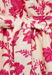 Robe florale avec une base crème, ornée de grands motifs de fleurs roses et magenta. Tissu texturé noué avec une ceinture assortie à la taille.