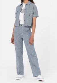 Krátký rukáv, zkrácená košile a kalhoty s vysokým pasem v modro-bílých vertikálních pruzích; má kapsy na hrudi a logo. Obléknuto s bílými teniskami.