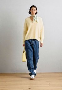 Cremfarbener V-Ausschnitt-Pullover, lockere blaue Jeans, weiße Sneaker und eine gelbe Tasche. Ein hellgrüner Schal, der um den Hals gebunden ist, verleiht einen sanften Akzent.