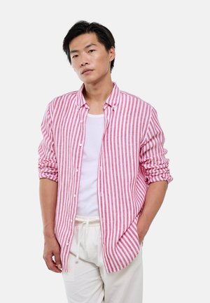 Jeune homme portant une chemise rayée verticalement rouge et blanche sur un t-shirt blanc avec un pantalon blanc, debout avec une main dans la poche.