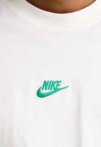 Camiseta blanca de algodón con cuello redondo. Presenta un logotipo de Nike bordado en verde en el pecho. Textura suave y lisa.