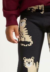 Svarta leggings med bruna tigertryck, inklusive detaljerade ansikten och ränder. Midjebandet är enkelt, och tyget verkar mjukt och elastiskt.
