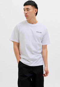 Valkoinen lyhythihainen T-paita pyöreällä kaula-aukolla, jossa on pieni musta "JACK & JONES" -logo rinnassa, yhdistettynä mustiin housuihin.