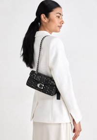 Kvinna i vit texturerad blazer bär en svart kviltad läderaxelväska med silvernitar och ett metallspänne, vänd åt höger.