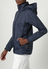 Morska granatowa wodoodporna kurtka z kapturem, zapinana na zamek błyskawiczny, z kieszeniami. Posiada teksturowane mankiety i logo na piersi. Gładki, lekki materiał.