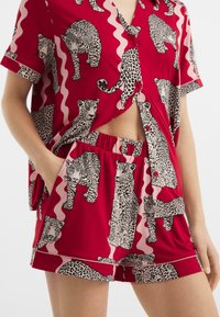 Rotes Oberteil mit kurzen Ärmeln und Shorts-Set mit Leopardenmuster und pinken Wellenmustern. Der Stoff ist glatt; das Design enthält Knopfdetails.