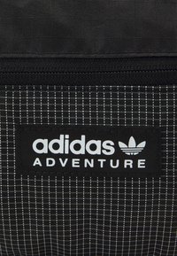 Svart tyg med ett vitt rutmönster och en sydd svart etikett med texten "adidas ADVENTURE" bredvid Adidas trefoil-logotyp.