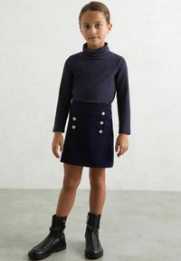 Námornícky dlhý rukáv turtleneck v kombinácii s ladiacou sukňou s zlatými gombíkmi. Čierne členkové topánky s reťazovými akcentmi dopĺňajú vzhľad.