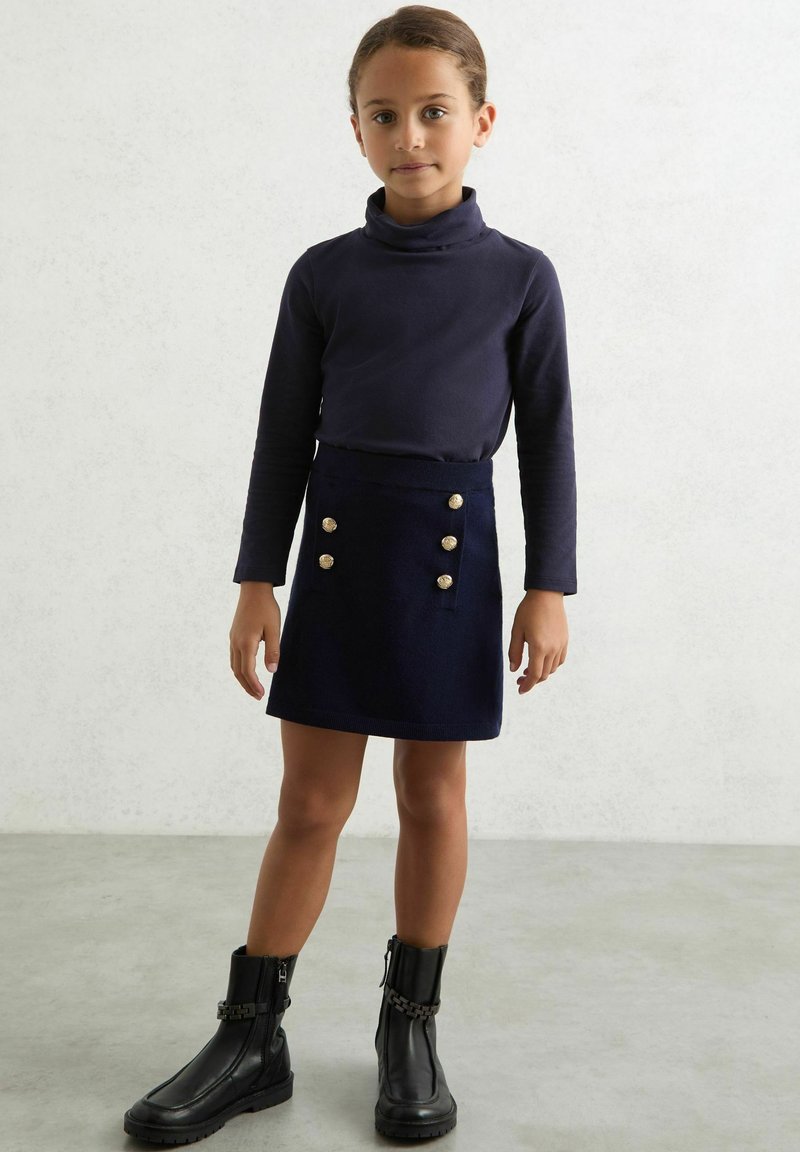 Námornícky dlhý rukáv turtleneck v kombinácii s ladiacou sukňou s zlatými gombíkmi. Čierne členkové topánky s reťazovými akcentmi dopĺňajú vzhľad.