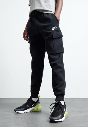 Personne portant un jogging cargo noir Nike avec des poignets élastiques et des baskets noires Nike avec des semelles jaune-vert, debout sur un sol gris.