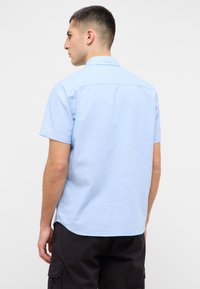 Camisa de manga corta azul claro con cuello, dobladillo redondeado y textura suave. La vista trasera muestra un diseño limpio y sencillo.