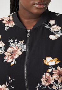 Černá květovaná bomber bunda s růžovými a žlutými květy, předním zipem a texturovanou tkaninou. Hladký límec doplňuje design.
